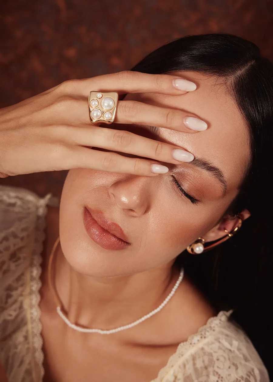Modelo com a mão sobre o rosto usando um anel dourado com pérolas